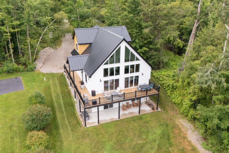 Drone view of a white lakeside farmhouse with a wraparound deck, surrounded by woods