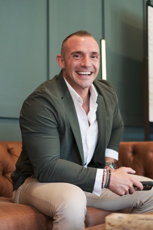 Scott Chain laughing, seated on a leather sofa in a green blazer